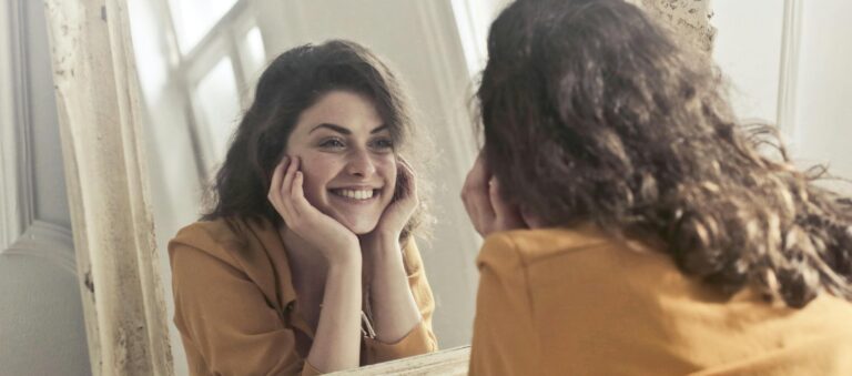 A cheerful woman smiles at her reflection in a vintage-style mirror, exuding positivity and warmth.