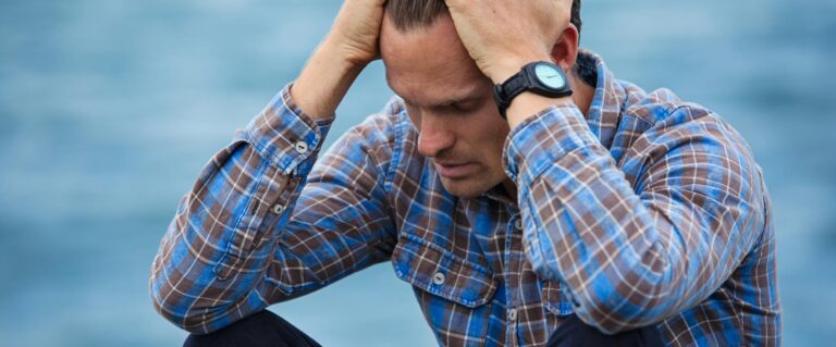 A man in a plaid shirt sits by the water looking distressed, symbolizing stress.