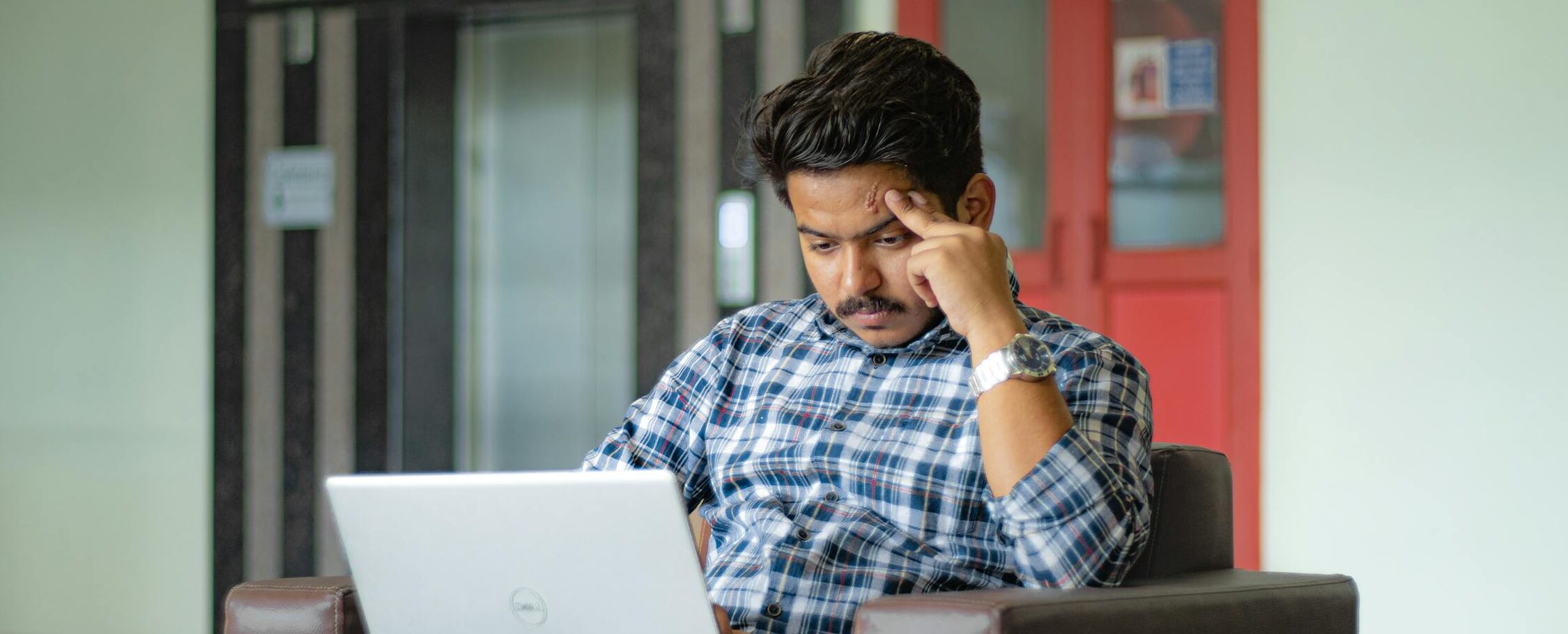 Man in plaid shirt concentrating on laptop work in a modern indoor setting.