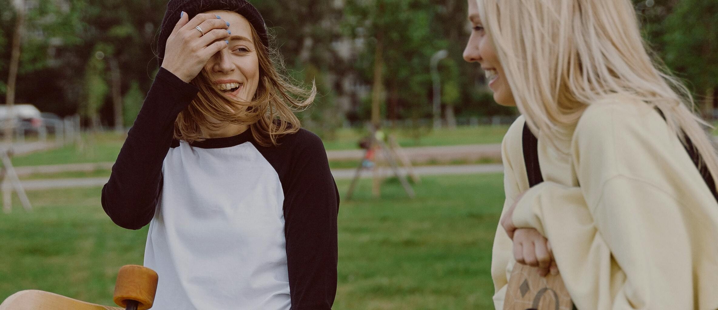 Two teenage girls laughing and holding longboards in a park, enjoying leisure time together.