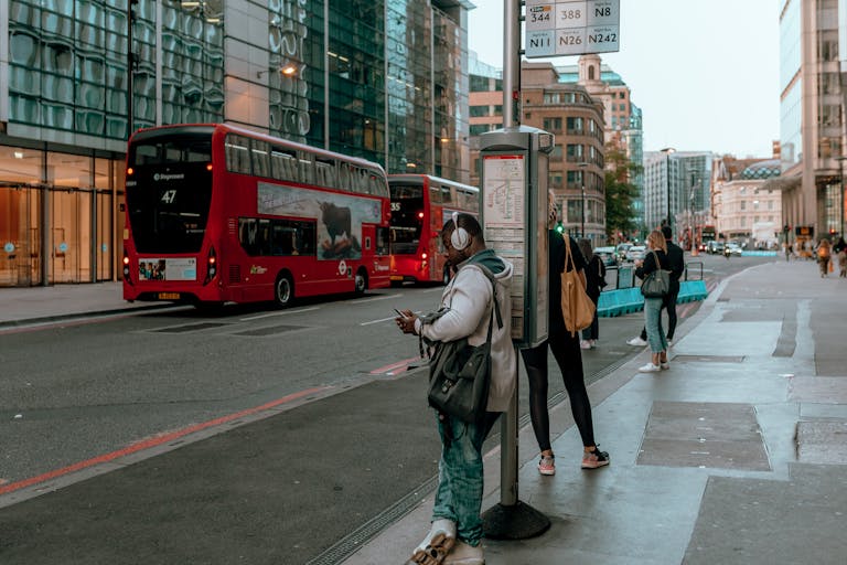 A busy street with people waiting at a bus stop and red double-decker buses passing by.