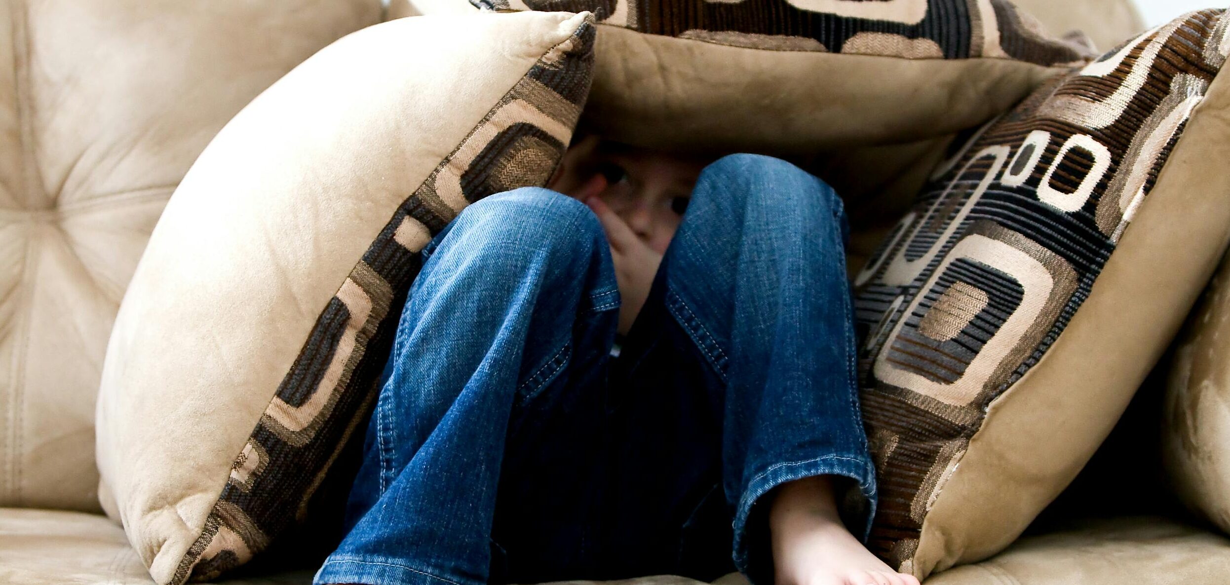 A child crouched under cushions on a couch, conveying a sense of playfulness or fear.