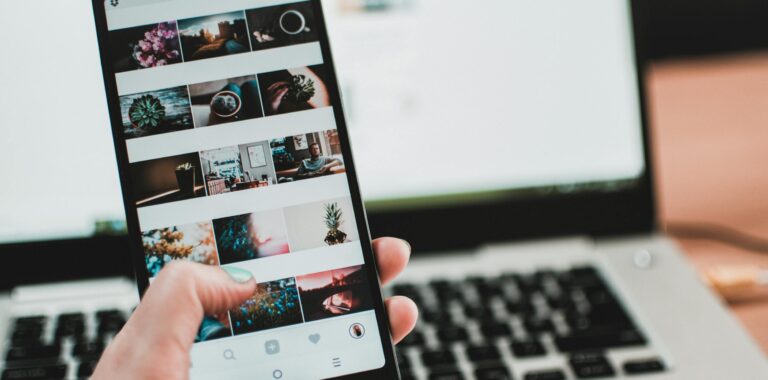 A person uses a smartphone to browse social media in front of a laptop, emphasizing modern technology and lifestyle.