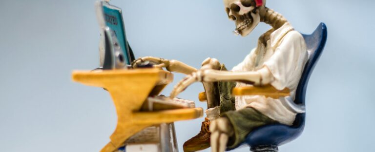 A quirky skeleton figurine wearing headphones sits at a desk with a computer, symbolizing burnout.