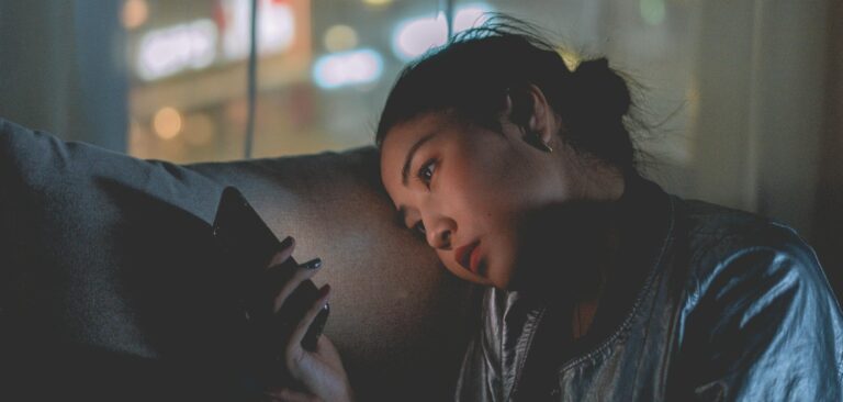 A woman looking at her mobile phone at night with city lights blurred in the background.