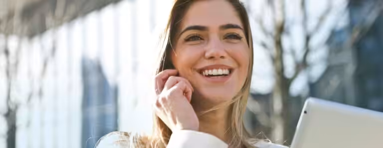 Confident businesswoman using her tablet and phone, smiling outdoors in sunlight.