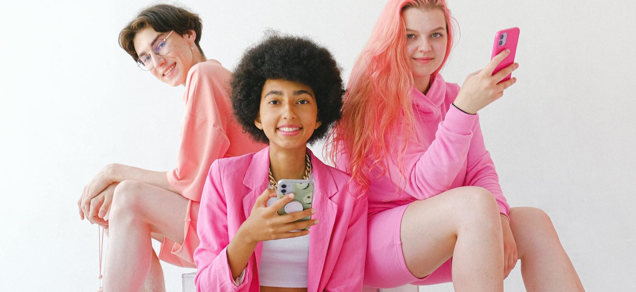 Group of teens in pink outfits using smartphones against a white background, exuding a modern and cheerful vibe.