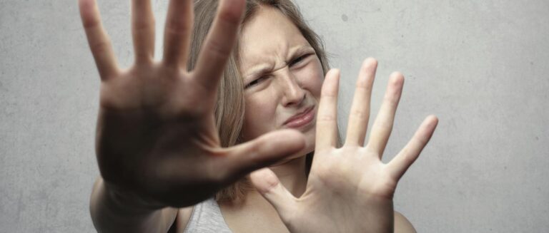 Portrait of a fearful woman in a gray tank top with hands pushed forward against a gray background.