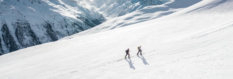 Two skiers climbing a sunlit snowy mountain slope in Ischgl, Austria, during winter.