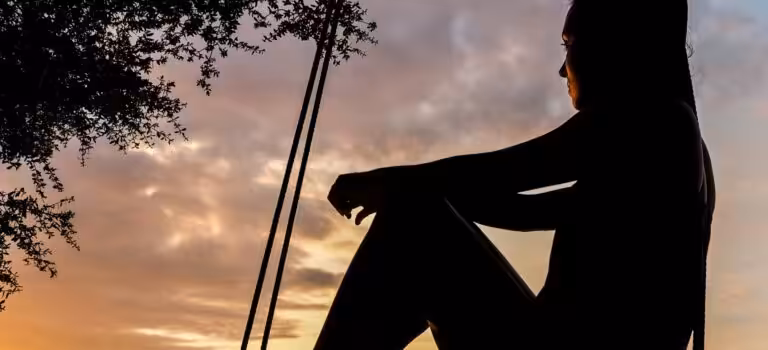 A serene sunset silhouette of a woman sitting on a swing by the seashore, evoking tranquility and reflection.