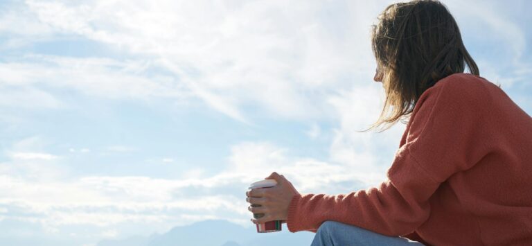 A woman in casual attire enjoys a hot drink while gazing at the sea, epitomizing relaxation.