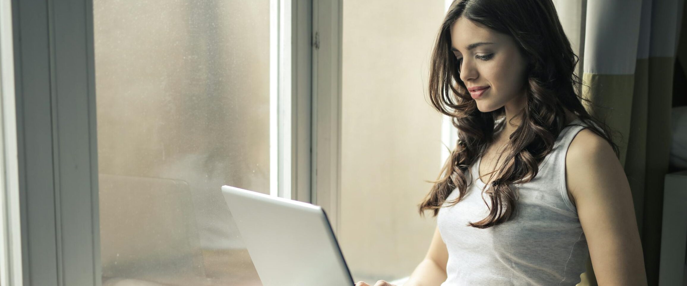 Adult woman sitting by window working remotely on a laptop indoors.