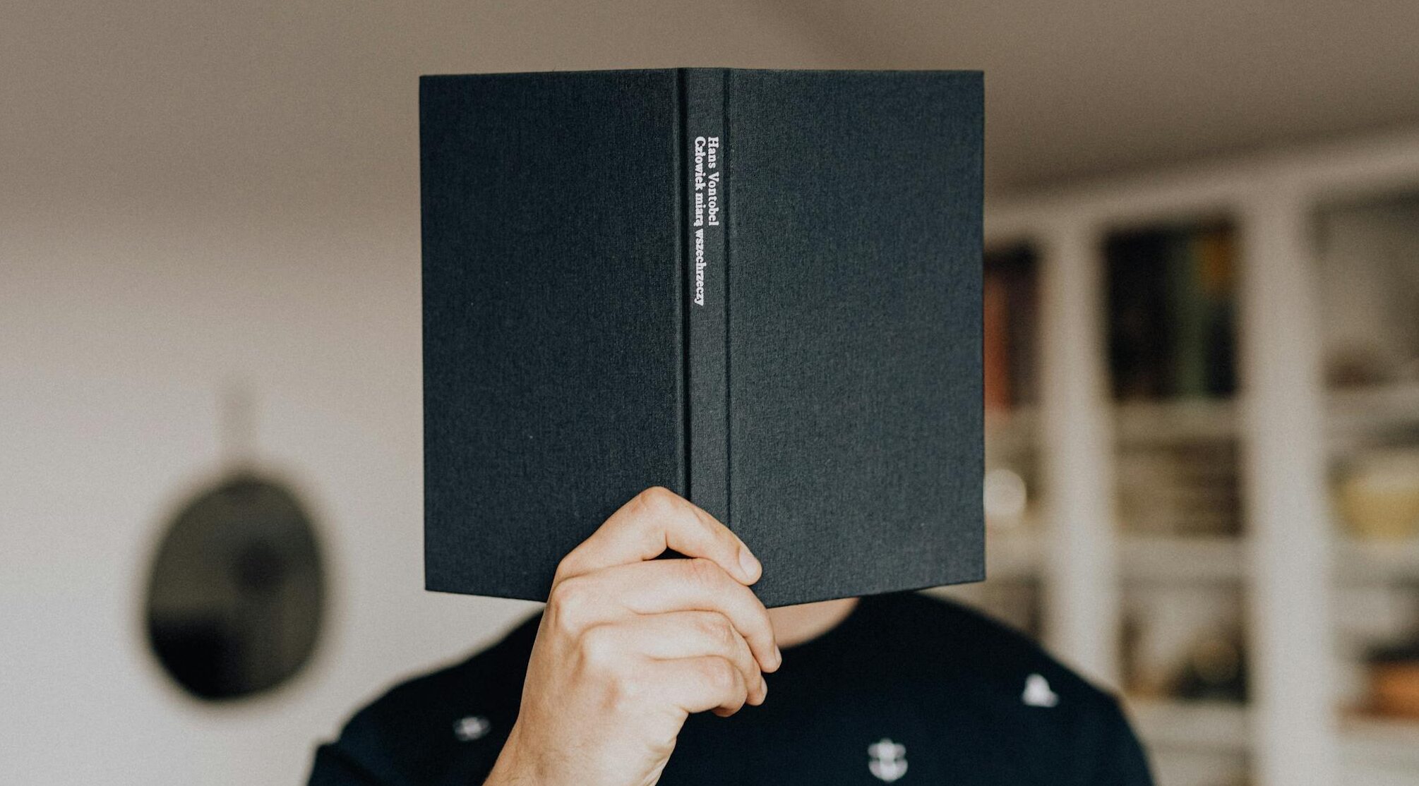 Faceless male holding opened black book in hand covering face while standing in middle of light room